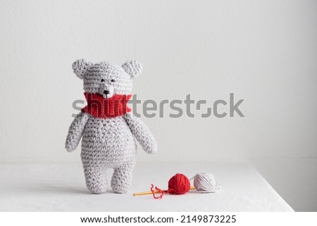 Crochet knitting cute gray, white, polar teddy bear with a red scarf stands on the table, homemade amigurumi with knitting hook and yarn