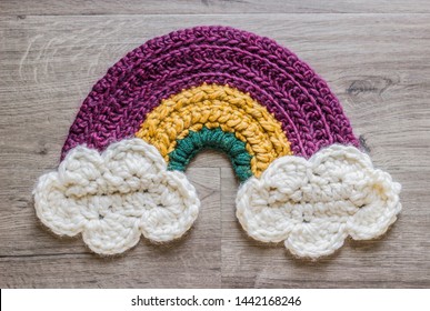 A Crochet Rainbow And Clouds Rests Against A Grey Wooden Backdrop.