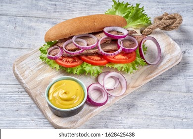 Crock Pot Italian Beef Sandwich Of Hoagie Bread Rolls With Beef And Vegetables: Tomatoes, Red Onion Rings, Fresh Lettuce Leaves, Served With Mustard And Barbeque Sauce, On A White Wooden Table