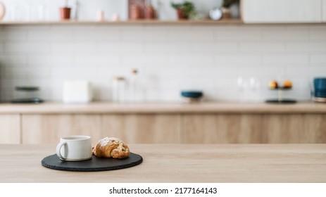 Croissant y café en la encimera de la cocina, contra el interior minimalista borroso con mobiliario moderno. Esfuerzo selectivo en pasteles caseros y bebida de té en taza sobre mesa de madera, espacio de copias, banner web