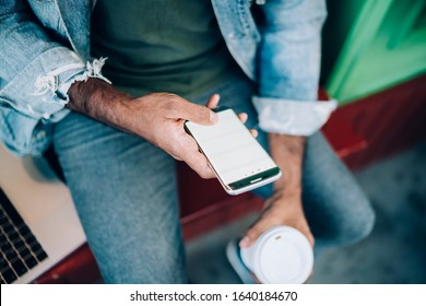 Crop From Above Male In Denim Sitting With Disposable Cup Of Coffee In Hand And Scrolling News Feed  On Social Media On Mobile Phone