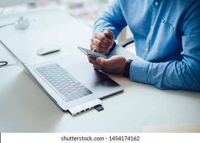 Crop From Above Of Modern Man In Blue Shirt Sitting At Working Table With Laptop In Modern Office And Using Mobile Phone