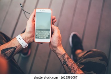 Crop from above view of casual man in camouflage pants and with tattooed arm messaging with mate using smartphone relaxing in outdoors of cafe