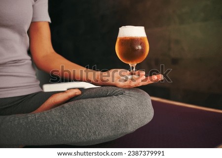 Crop anonymous barefoot female in activewear meditating with glass of fresh foamy beer while sitting on mat in light room