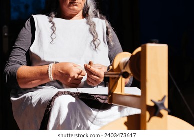Crop of anonymous concentrated elderly female in casual clothes sitting while using wooden spinning machine to make thread on sunny day against dark background - Powered by Shutterstock