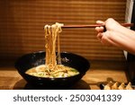 Crop anonymous female with chopsticks eating yummy ramen soup with noodles and meat in black bowl at ramen restaurant.