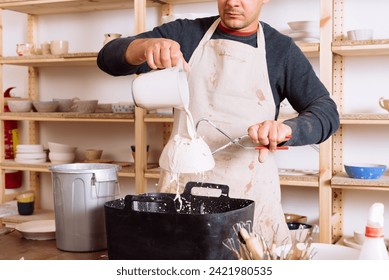 Crop of anonymous male artisan in apron pouring ceramic glaze over plate while working at pottery workshop against background of shelf with ceramics - Powered by Shutterstock