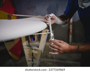 Crop anonymous male master in respirator measuring thickness of surfboard with ruler while shaping board placed in racks in workshop - Powered by Shutterstock