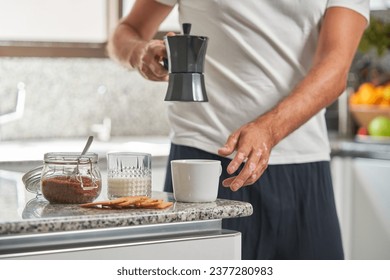 Crop anonymous man pouring fresh coffee from geyser coffeemaker in ceramic cup placed on counter near glass of milk and jar with brown sugar while preparing breakfast