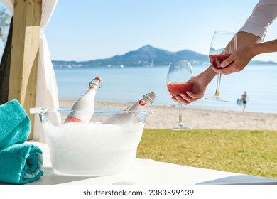 Crop anonymous person putting pair of goblets with rose champagne on canopy bed placed near sea during romantic date on beach in summer day
