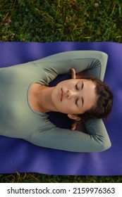 Crop of asian female woman closing eyes lying down on yoga mat among grass. Above view of relaxing girl in blue wear resting peacefully outdoors after doing yoga exercises. Concept of inner harmony.