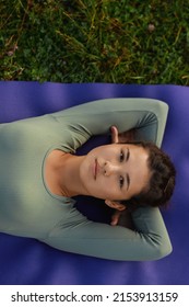 Crop Of Asian Female Woman Lying Down On Yoga Mat In Nature. Top View Of Relaxing Girl Looking At Camera In Blue Wear Resting Among Grass After Doing Yoga Exercises. Concept Of Inner Harmony.