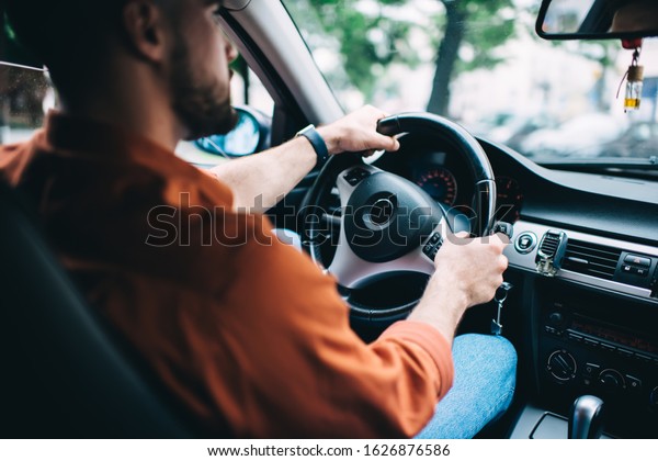 Crop back view of bearded male driver in
casual clothing sitting in modern car and turning steering wheel
driving on blurred
background