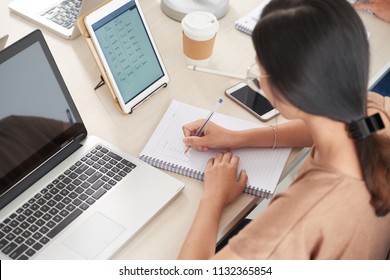 Crop back view of brunette woman in casual clothes sitting at light wooden desk with tablet and laptop and writing into notepad 