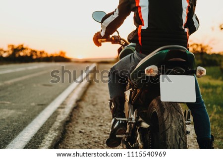 Crop back view of male rider in protective boots and jacket moving on highway from roadside on backlit blurred background of empty route