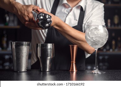 Crop of bartender's hands adding pressed citrus juice into stainless steel cocktail shaker. Measuring cup and wine glass filled with ice cubes on right side