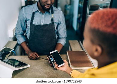 Crop Black Cashier Man Servicing Woman In Yellow Dress With Contactless Payment Using Mobile Phone And Payment Terminal In Cafe Standing At Counter