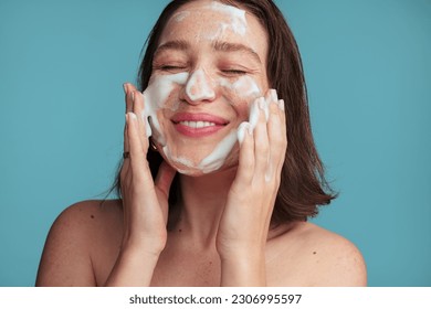 Crop cheerful young woman with dark hair smiling happily with closed eyes while cleaning face with foam during skincare procedure in blue studio - Powered by Shutterstock