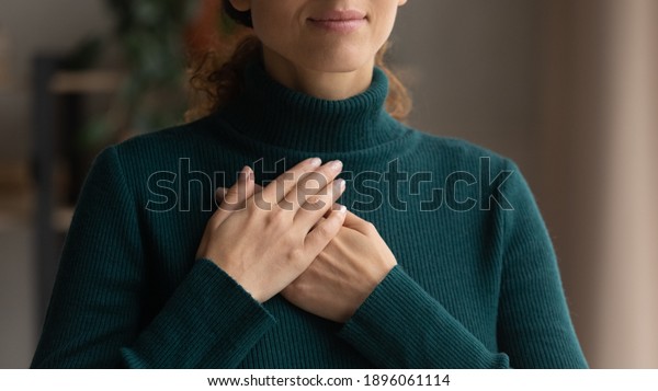 Crop close up of Caucasian woman hold hands in\
prayer at heart chest feel religious superstitious. Young female\
believer being grateful thankful pray to God. Faith, belief,\
religion concept.