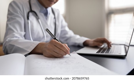 Crop close up Indian woman doctor in white uniform with stethoscope taking notes, using laptop, writing in medical journal, professional therapist practitioner filling documents or patient card - Powered by Shutterstock