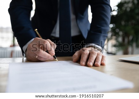 Crop close up of male CEO or boss in suit stand in office put signature on paper document making agreement. Businessman sign paperwork close business financial deal at workplace. Legislation concept.