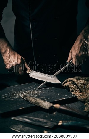 Crop faceless male chef sharpening metal blade of knife with sharpener while working in dark kitchen