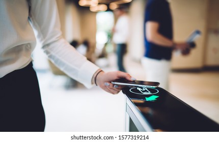 Crop female in trendy formal clothes swiping smartphone while passing through automatic access control in modern foyer of business center on blurred background