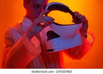 Crop Focused Female In Transparent Coat Putting On Modern VR Goggles While Standing In Bright Studio With Neon Lights Before Exploring Cyberspace