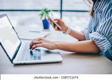 Crop hands of faceless female office employee in casual clothing typing on keyboard of empty screen laptop and surfing on mobile phone at modern office - Shutterstock ID 1589711674