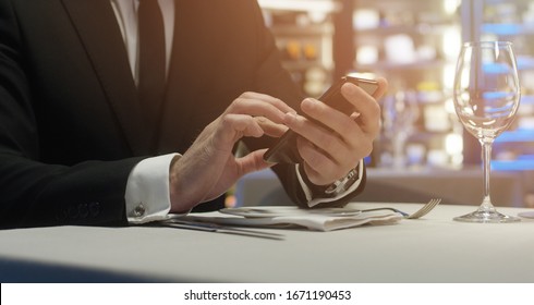 Crop Of Male Using Smartphone At Table In Luxury Restaurant. Close Up Of Businessman Waiting For Lunch With Colleague And Typing, Writing Message On Call Phone. Concept Of Gadgets.