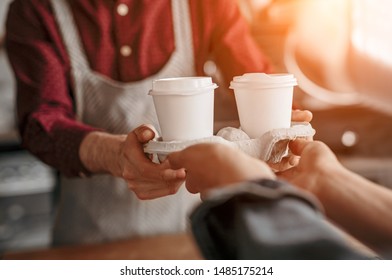 Crop Man In Apron Giving Disposable Cups Of Hot Coffee In Carton Holder To Customer In Bright Morning Sunrays