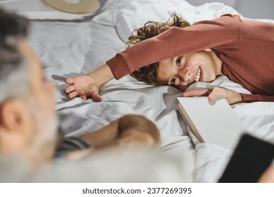 Crop man and smiling boy in sleepwear with book relaxing together on bed in cozy bedroom at home while looking at each other