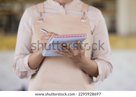 Crop midsection close up view, unknown waitress in apron hold pen and notepad ready to take order standing at workplace. Good customer services, food and drink commercial establishment worker workflow