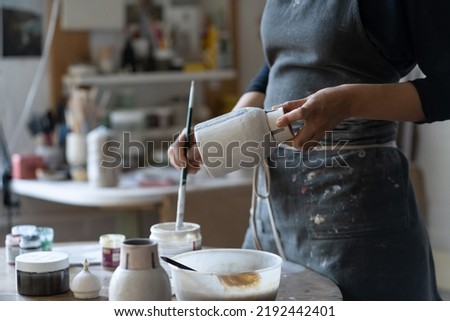 Crop photo of woman artisan making earthenware vase. Female skilled worker hands prepare clay item with brush for painting in big workroom. Young craftswoman enjoys creating things from clay closeup