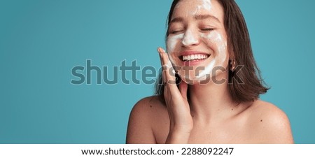 Crop positive young female millennial with brown hair smiling with closed eyes while washing face with foam cleanser against blue background