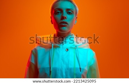 Crop self assured young female millennial with shirt blond hair in transparent raincoat looking at camera against orange background in neon studio