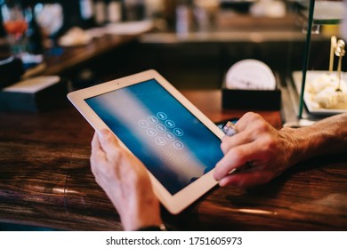 Crop seniour client using tablet for entering graphic key for paying order while leaning on wooden bar counter