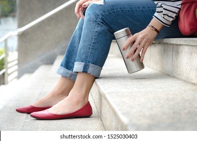Crop shot of an attractive woman wearing jeans with stylish ruby red flat shoes sitting on a stairs, her hand grab a reusable isolated water bottle. Plastic-free, No straw, Eco, Zero waste concept.