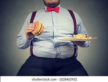 Crop shot of big man in formal clothes sitting and consuming plate with fast food on gray. 