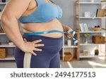 Crop shot overweight, obese woman in sports bra holding measuring tape around fat belly to check how much weight she has gained and how many centimeters she has added in her waist. Fat body concept