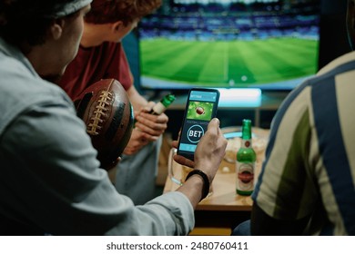 Crop shot of young male friends betting on team in app on smartphone while watching football match on TV - Powered by Shutterstock