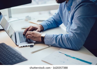 Crop side view of man in blue shirt sitting at white desk with paper charts and typing while working on laptop  - Shutterstock ID 1620357388