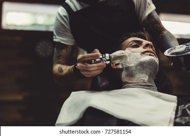 Crop stylish applying foam on customer's cheeks for shaving while working in barbershop.
