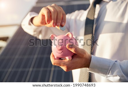 Crop unrecognizable businessman inserting coin into piggy bank while standing against blurred photovoltaic solar panel as concept of cheap alternative energy
