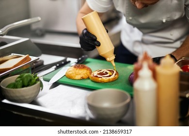 Crop View Of Chef's Hands Pouring Sauce On Cheeseburger In Restaurant. Male Hands In Gloves Preparing Tasty Beef Burger Inside Professional Kitchen. Concept Of Food Cooking.