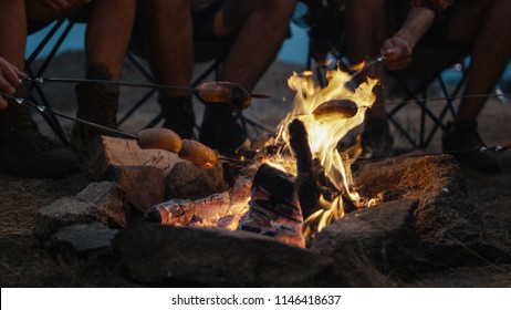 Crop view of people gathering around burning campfire and frying sausages on skewers in bright flame making dinner