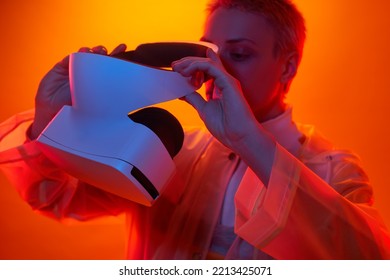 Crop Young Futuristic Woman With Short Blond Hair In Transparent Raincoat Wearing Modern VR Headset Against Orange Background In Neon Studio