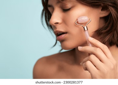 Crop young Hispanic female with dark hair and perfect skin massaging face with pink jade roller during beauty treatment against blue background