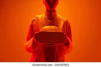 Crop Young Woman In Futuristic Clothes Carrying Hi Tech VR Goggles Under Bright Orange Neon Illumination