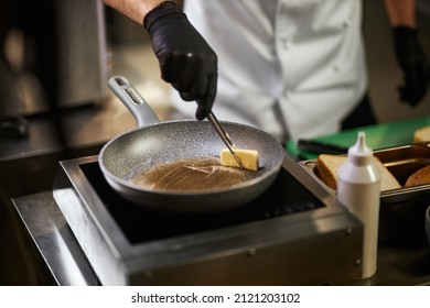Crope view of chef's hands in black gloves melting piece of butter in frying pan. Male in chef uniform using tweezers for cooking with ingredients and equipment on background. Concept of cooking. 